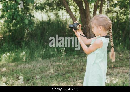 Ein glückliches kleines sieben Jahre altes Mädchen fotografiert eine Sommer-Naturlandschaft mit einer Kamera mit Live-Ansicht. Kinder adoptieren ihre Eltern Hobbys. Summe Stockfoto