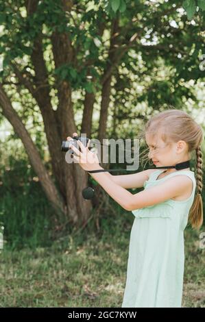 Ein glückliches kleines sieben Jahre altes Mädchen fotografiert eine Sommer-Naturlandschaft mit einer Kamera mit Live-Ansicht. Kinder adoptieren ihre Eltern Hobbys. Summe Stockfoto
