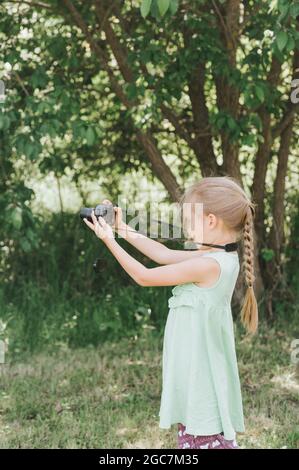 Ein glückliches kleines sieben Jahre altes Mädchen fotografiert eine Sommer-Naturlandschaft mit einer Kamera mit Live-Ansicht. Kinder adoptieren ihre Eltern Hobbys. Summe Stockfoto