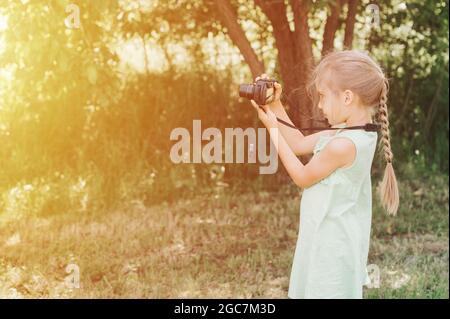 Ein glückliches kleines sieben Jahre altes Mädchen fotografiert eine Sommer-Naturlandschaft mit einer Kamera mit Live-Ansicht. Kinder adoptieren ihre Eltern Hobbys. Summe Stockfoto