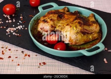 Gegrilltes Hähnchen mit Tomaten. Gebratenes Huhn in einem blauen Keramikblech, auf einem schwarzen Blech Stockfoto