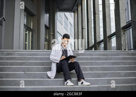 Vorderansicht eines jungen indischen männlichen Arztes, der auf der Zwischenablage schrieb und auf der Treppe vor dem modernen Krankenhaus saß. Fokussierter Arzt, der Arbeitsunterlagen, Versicherungsunterlagen oder Patientenunterlagen ausfüllt Stockfoto