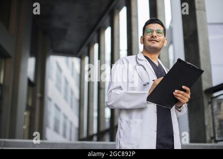Nahaufnahme Porträt von pensive professionelle männliche arabische Arzt schreiben RX Rezept auf Zwischenablage. Der männliche Arzt füllt die medizinische Form aus, die im Freien der modernen Klinik steht. Speicherplatz kopieren Stockfoto