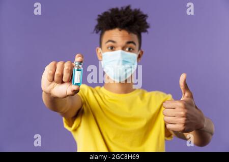 Nahaufnahme des aufrührenden jungen hispanischen Mannes in der medizinischen Maske, die Ampulle mit einem Kovid19-Impfstoff hält, Coronavirus-Medikament, zeigt Daumen hoch und wird angeschossen Stockfoto