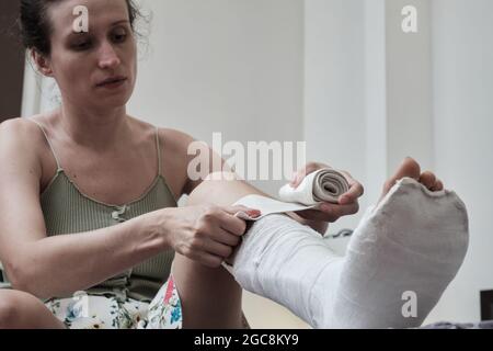Eine Frau, die auf einem Bett sitzt, legt einen elastischen Verband auf ein gebrochenes Bein. Das Bein ist in einem Gipsabguss. Heimrehabilitation nach einem gebrochenen Bein. Hochwertige Pho Stockfoto