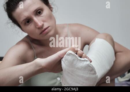 Frau sitzt auf dem Bett und hält ein gebrochenes Bein. Das Bein ist in einem Gipsabguss. Heimrehabilitation. Hochwertige Fotos. Stockfoto