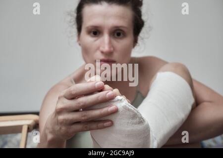 Frau sitzt auf dem Bett und hält ein gebrochenes Bein. Das Bein ist in einem Gipsabguss. Heimrehabilitation. Hochwertige Fotos. Stockfoto