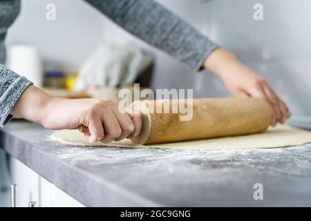 Nahaufnahme der Hände einer unbekannten kaukasischen Frau, die Teigpaste für Croissants mithilfe von Nudelholz auf der Küchentische zu Hause zubereitet - Hausmannskost c Stockfoto