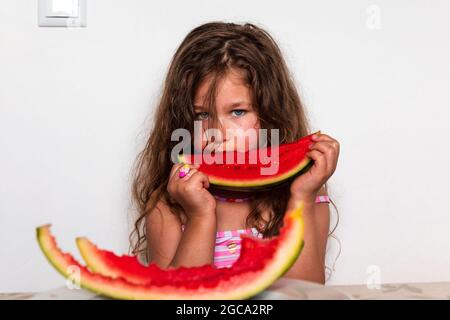 Kaukasisches Mädchen sitzt am Tisch und isst Wassermelone. Gebräuntes kleines Mädchen mit blauen Augen schaut in die Kamera und hält ein Stück Wassermelone in den Händen. Je zwei Stockfoto