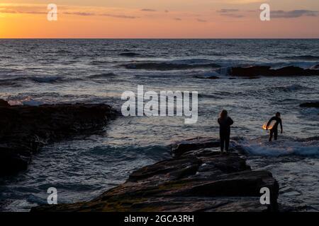 Sonnenuntergang am Strand von Trebarwith Strand, North Cornwall, mit einer jungen Frau in Silhouette, die einen jungen Surfer aus dem Wasser auftaucht. Stockfoto