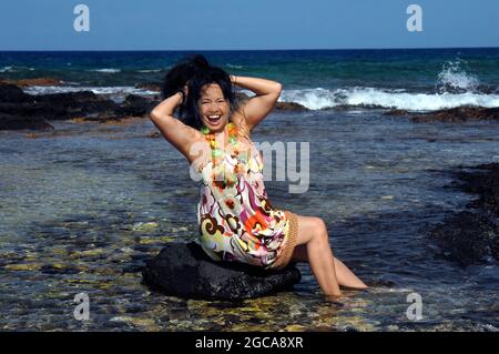 Hawaiianerin sitzt auf einem Felsen im flachen Wasser und feiert, indem sie ihre Haare hochwirft und posiert. Anaehomalu Bay an der Kohala Küste der Big Isla Stockfoto