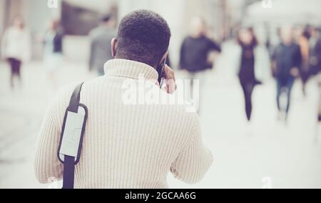 Schwarzer Mann aus der Generation der Millennials geht die Straße im Stadtzentrum entlang und spricht auf einem Smartphone. Rückansicht. Stockfoto
