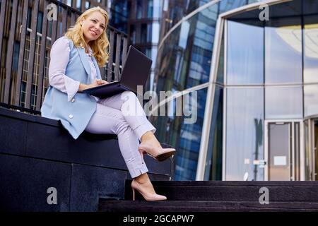 Lächelnde Frau im Business-Anzug mit Laptop sitzt im Freien. Stockfoto