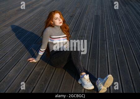 Trauriges Rotschopf-Mädchen sitzt allein an einem sonnigen Tag auf einem hölzernen Hintergrund im Park. Junge Frau allein. Traurigkeit und Depression Konzept Stockfoto