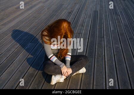 Trauriges Rotschopf-Mädchen sitzt allein an einem sonnigen Tag auf einem hölzernen Hintergrund im Park. Junge Frau allein. Traurigkeit und Depression Konzept Stockfoto