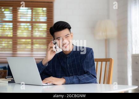Junge asiatische Mann telefonieren und arbeiten von zu Hause mit Laptop-Computer, freiberuflich mit Notebook ins Internet online und sprechen auf Smartphone, inländische l Stockfoto