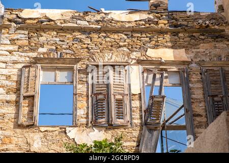 Beschädigtes Steinwandgebäude, kaputte alte hölzerne Fensterläden und Fenster mit Blick auf den blauen Himmel. Verlassene alte Verwesung Haus ruiniert vernachlässigte Struktur bei Syros Stockfoto