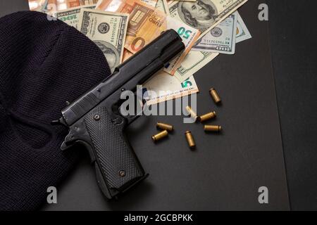 Kriminelles Geld. Pistole und Sturmhaube auf Dollar- und Euro-Banknoten, schwarzer Hintergrund, Draufsicht. Bewaffneter Raub, Einbruchskonzept, Stockfoto