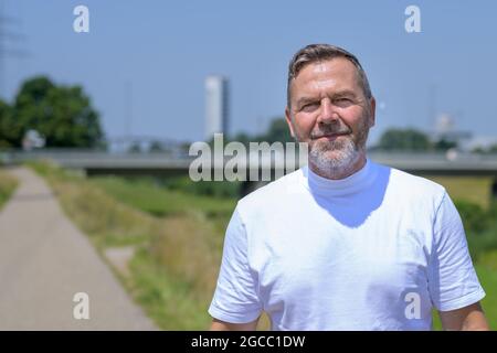 Nahaufnahme eines bärtigen Mannes mittleren Alters, der die Kamera anlächelt, während er an einem schönen sonnigen Sommertag auf einem Fußweg neben einem Fluss joggt Stockfoto