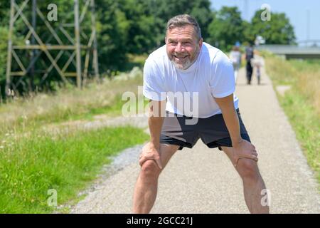 Ein glücklicher, gesunder Mann, der sich beim Joggen auf den Knien nach vorne lehnt, während er mit einem strahlenden Lächeln in Gesundheit, Fitness und Tat nach vorne blickt Stockfoto