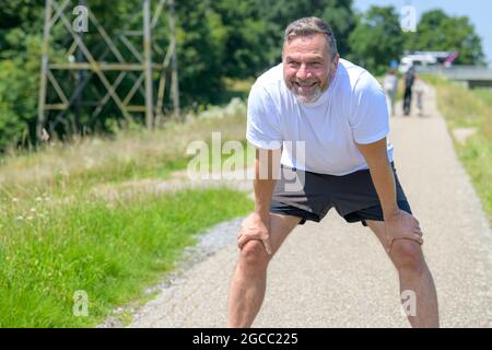 Ein glücklicher, gesunder Mann, der sich beim Joggen auf den Knien nach vorne lehnt, während er mit einem strahlenden Lächeln in Gesundheit, Fitness und Tat nach vorne blickt Stockfoto