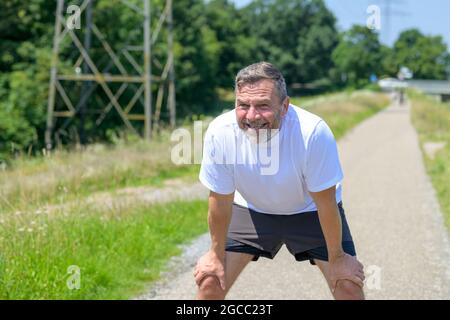 Erschöpfter, gesunder Mann, der sich ausruht, während er joggt und sich auf den Knien nach vorne lehnt, während er nach vorne blickt Stockfoto