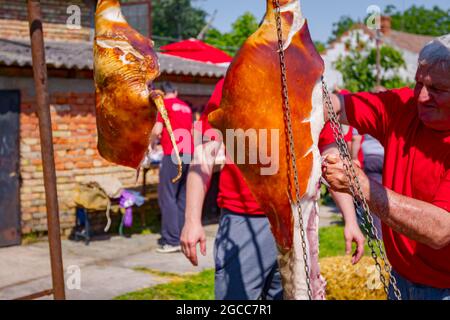 Aradac, Vojvodina, Serbien - 21. Mai 2016: Zwei rohe Schweineschinken hängen oben im Dorfhof, Metzger schneidet Fleisch. Stockfoto