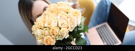 Frau hält einen großen Strauß Rosen und ein Glas Wein Auf ihrem Schoß ist ein Laptop Stockfoto