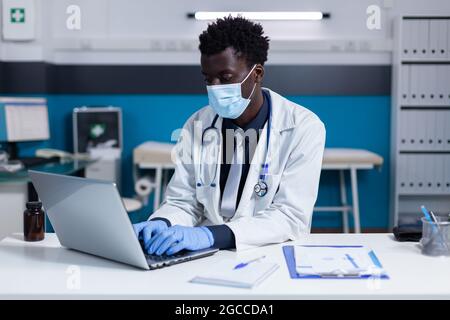 Schwarzer Mann mit Arzt Beschäftigung mit Laptop auf dem Schreibtisch für Medizin Rezept in der Arztpraxis. Der afroamerikanische Spezialist für das Gesundheitswesen tippt auf der Tastatur, während er eine Gesichtsmaske trägt Stockfoto
