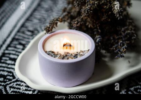 Duftkerze aus Lavendel mit Holzdocht und getrockneten Kräutern auf weißem Keramikblech Stockfoto