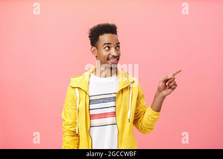Innenaufnahme eines faszinierten und erfreut afroamerikanischen männlichen Studenten in trendiger gelber Jacke, der mit Dreamy in die obere rechte Ecke zeigt und blickt Stockfoto