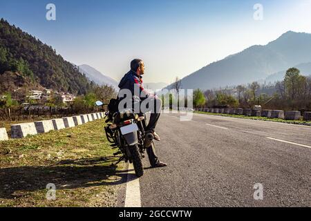 Mann, der am Motorrad auf einer asphaltierten Straße mit schöner natürlicher Aussicht am Morgen sitzt, wird am Shergaon arunachal pradesh india aufgenommen. Stockfoto