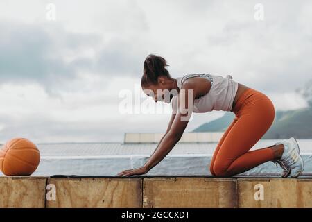 Seitenansicht einer lächelnden Frau beim Training im Freien mit einem Basketball vor der Tür. Sportlerin übt Yoga auf dem Dach. Stockfoto