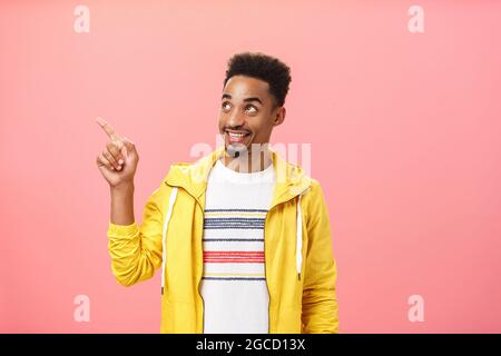 Aufnahme eines kreativen afroamerikanischen Mannes in der Taille mit Afro-Haarschnitt in einer trendigen gelben Jacke über dem T-Shirt, der auf die linke obere Ecke zeigt Stockfoto