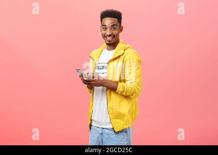 Stilvoller entzückter afroamerikanischer Mann mit afro lockigem Haarschnitt, der halb über die rosa Wand gedreht steht und das Smartphone in einer gelben trendigen Jacke hält Stockfoto