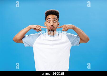 Ein verspielter, fröhlicher afroamerikanischer Kerl mit Tätowierung auf der Handfläche, der den Atem anhält und freudig auf die Wangen mit Zeigefinger stockt Stockfoto