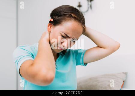 Kaukasische Frau, die zu Hause sitzt und unter Schmerzen im Nacken und in der Wirbelsäule leidet. Stockfoto