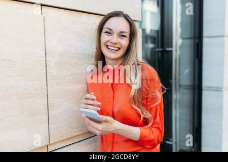 Eine kaukasische Frau hält ein Smartphone in den Händen und lacht über die Kamera in der Stadt Stockfoto