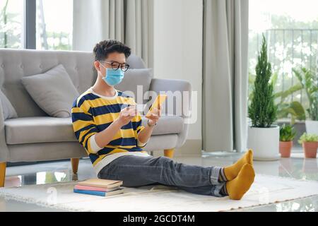 Junger glücklicher, gutaussehender Mann in medizinischer Maske sitzt am Boden und kauft online auf dem Smartphone. Stockfoto