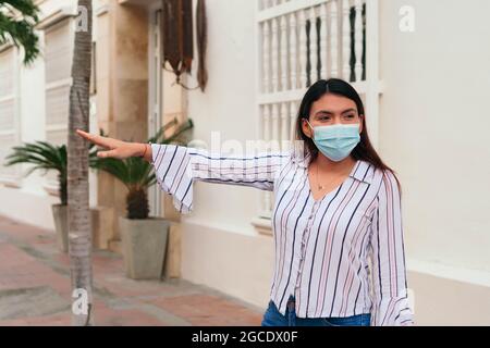 Frau mit Maske, die Einkaufstaschen in der Hand hält und lächelt, während sie ein Taxi anruft. Warten auf ein Taxi nach einem langen Einkaufstag. Stockfoto