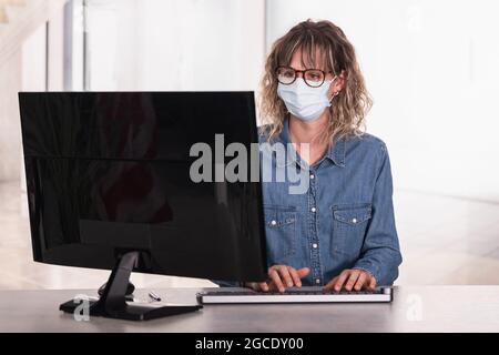 Erwachsene Frau trägt eine schützende Gesichtsmaske im Büro mit Computer Stockfoto