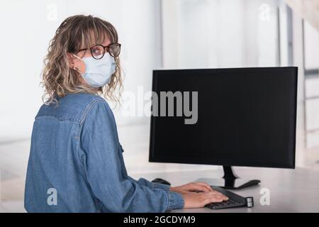Rückansicht einer erwachsenen Frau, die im Büro eine schützende Gesichtsmaske trägt und einen Computer mit dunklem Bildschirm verwendet, um Platz zu kopieren Stockfoto