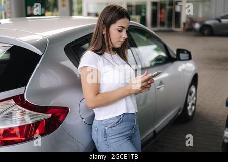 Attraktive junge Frau benutzte ihr Telefon und lehnte sich an der Tankstelle auf ihr Auto. Wartende Mitarbeiterin an der Tankstelle Stockfoto