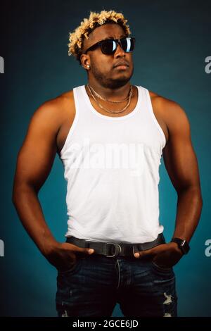 Dies ist ein Studioporträt eines Afrikaners in Sonnenbrillen und einem weißen T-Shirt auf dunkelblauem Hintergrund. Ein schwungvoller Kerl mit modischer Frisur und Zöpfen hält seine Hände in den Taschen und schaut weg. Werbeporträt. Stockfoto