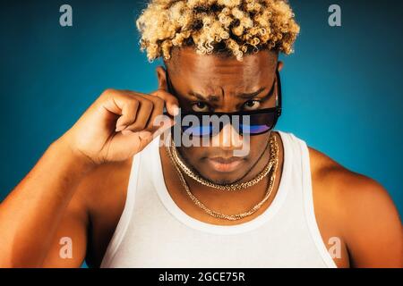 Dies ist ein Studioporträt eines Afrikaners in Sonnenbrillen und einem weißen T-Shirt auf dunkelblauem Hintergrund. Ein schwül getragener Kerl mit modischer Frisur und Zöpfen blickt direkt unter der Brille in die Kamera. Stockfoto