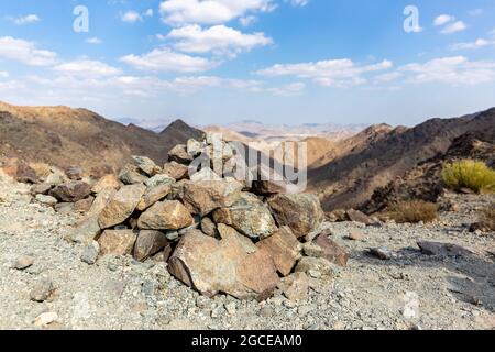 Cairn, ein vom Menschen gemachter Haufen (oder Stapel) aus Steinen, der die Bergspitze in Hatta, Haschar Mountains, markiert, mit trockenen, kargen Bergketten im Hintergrund. Stockfoto