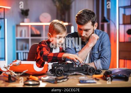 Fürsorglicher Vater lehrt seinen kleinen Sohn, zu Hause ein rotes Spielzeugauto zu reparieren. Kaukasischer Mann und Junge sitzen am Tisch und verwenden Lötkolben mit Schraubendreher. Stockfoto