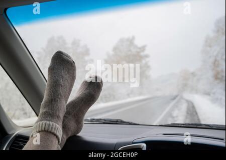 Weibliche Füße in Wollsocken auf dem Armaturenbrett eines Autos im Winter. Windschutzscheibenansicht Stockfoto