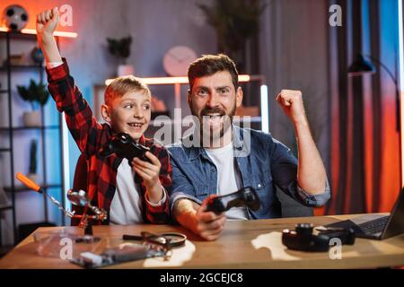 Kaukasischer Mann und Junge feiern den Sieg in Videospielen, während sie zusammen am Tisch sitzen. Junger Vater und kleiner Sohn gestikulieren emotional während der Unterhaltung zu Hause. Stockfoto