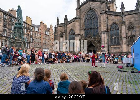 Edinburgh, Schottland, Großbritannien. August 2021. An einem sonnigen Sonntagnachmittag war die Royal Mile voll mit Besuchern, die auf der Suche nach der begrenzten Straßenunterhaltung waren, die während des viel zurückgehenkten Edinburgh Fringe Festivals in diesem Jahr geboten wurde. Zwei Bühnen sind für Darsteller vorgesehen und diese erwiesen sich den ganzen Tag über als beliebt. Bild: Die Menge beobachtet den Darsteller auf dem Parliament Square vor der St. Giles Cathedral. . Iain Masterton/Alamy Live Nachrichten. Stockfoto